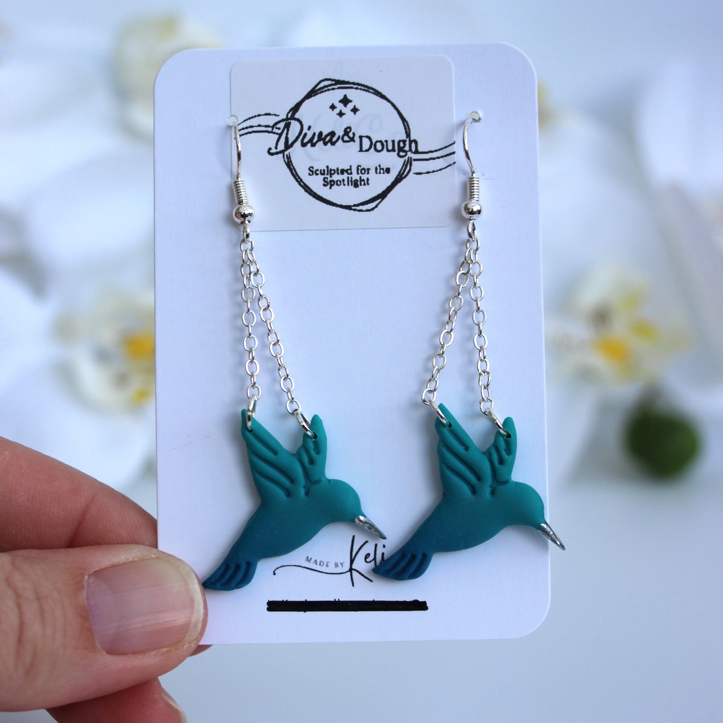 Teal hummingbird-shaped earrings on a branded card held by a hand with a blurred floral background