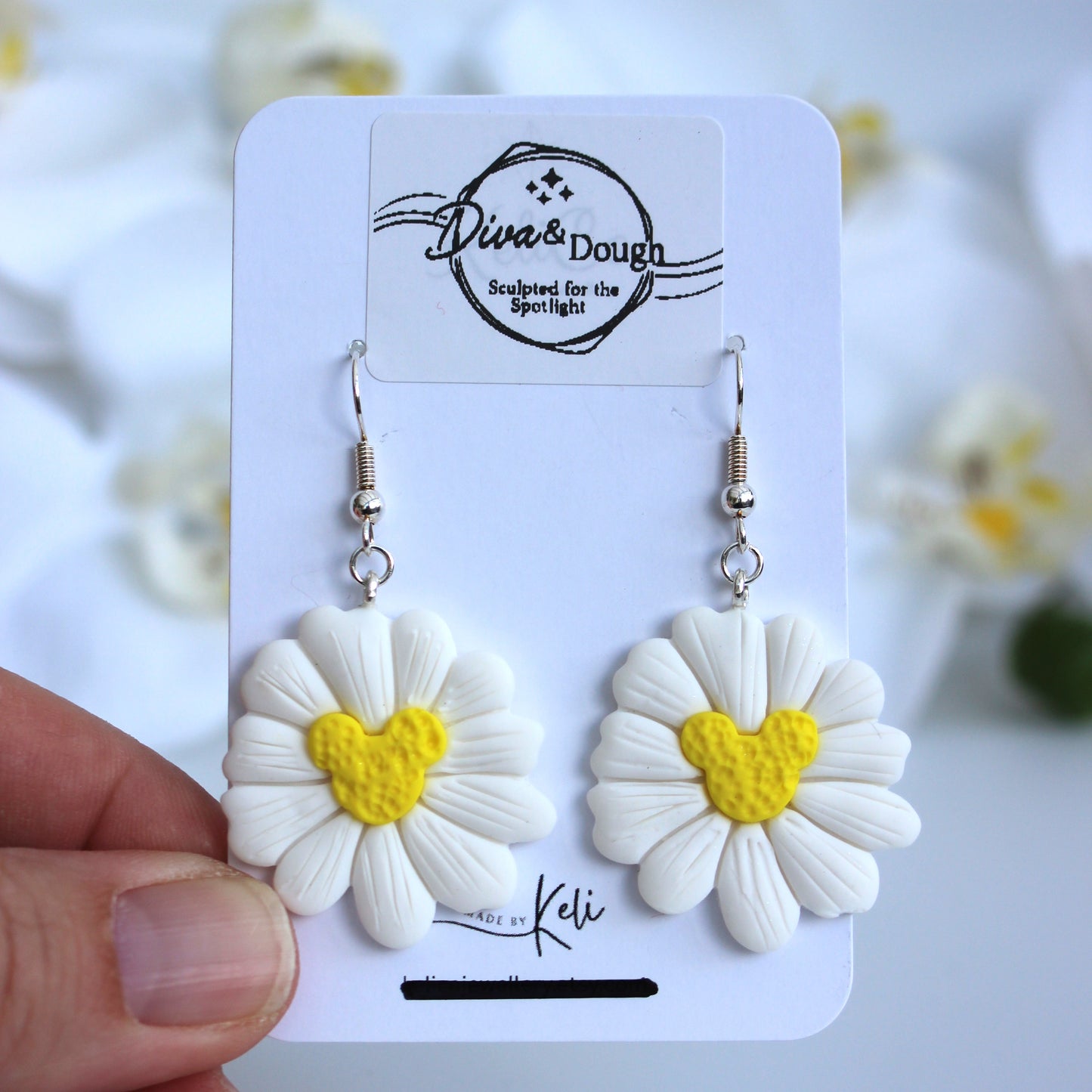 White flower-shaped earrings with a yellow mouse ears shaped center on a branded card held by a hand.