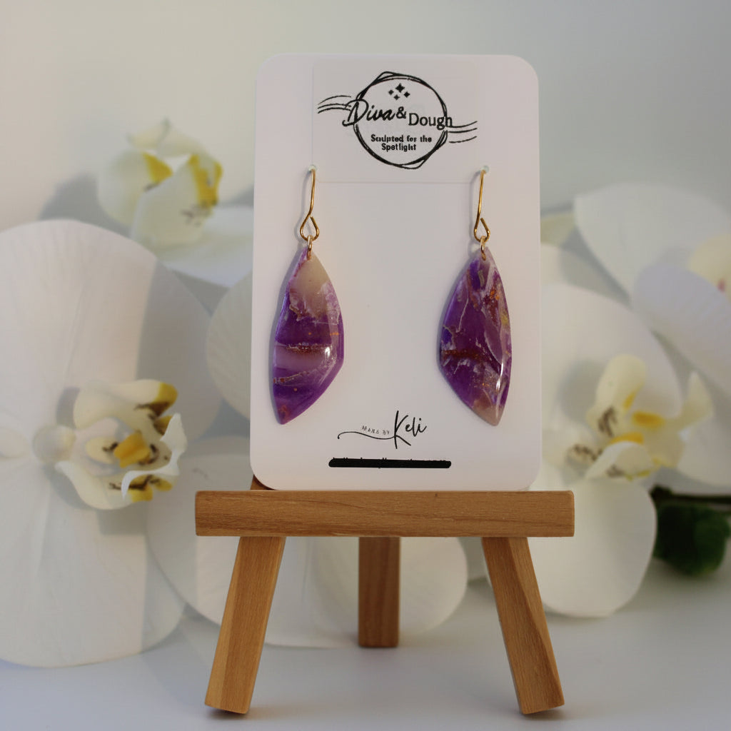 Purple teardrop earrings on a display card with a wooden stand, surrounded by white flowers.