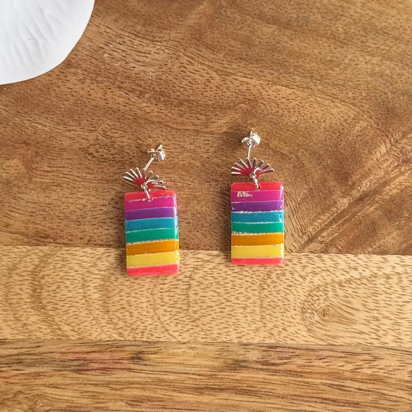 Pair of Diva & Dough hand-sculpted rectangular earrings with horizontal rainbow stripes and silver fan-shaped studs on a wooden background.