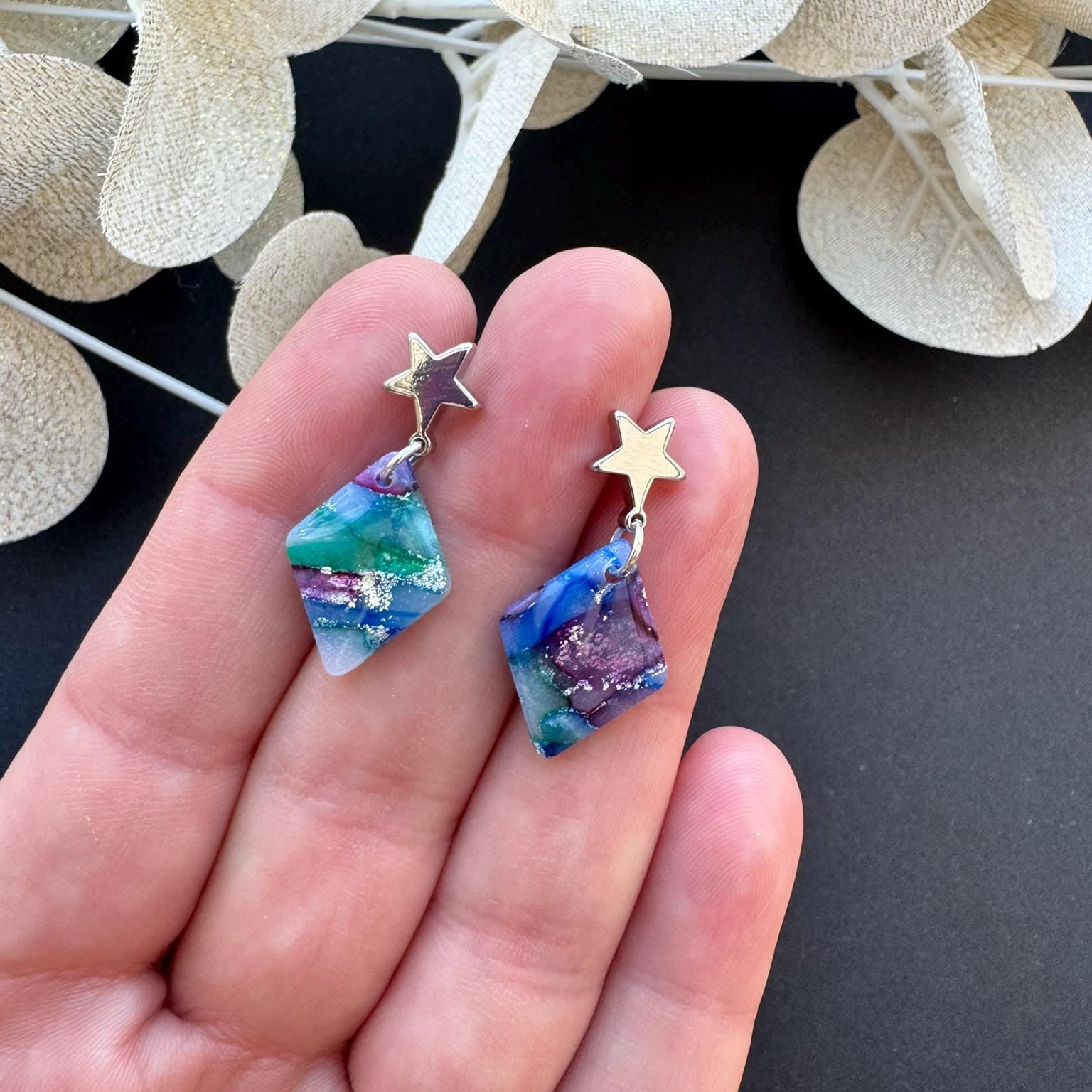 Colorful star-shaped earrings held in a hand with a dark background and decorative leaves.
