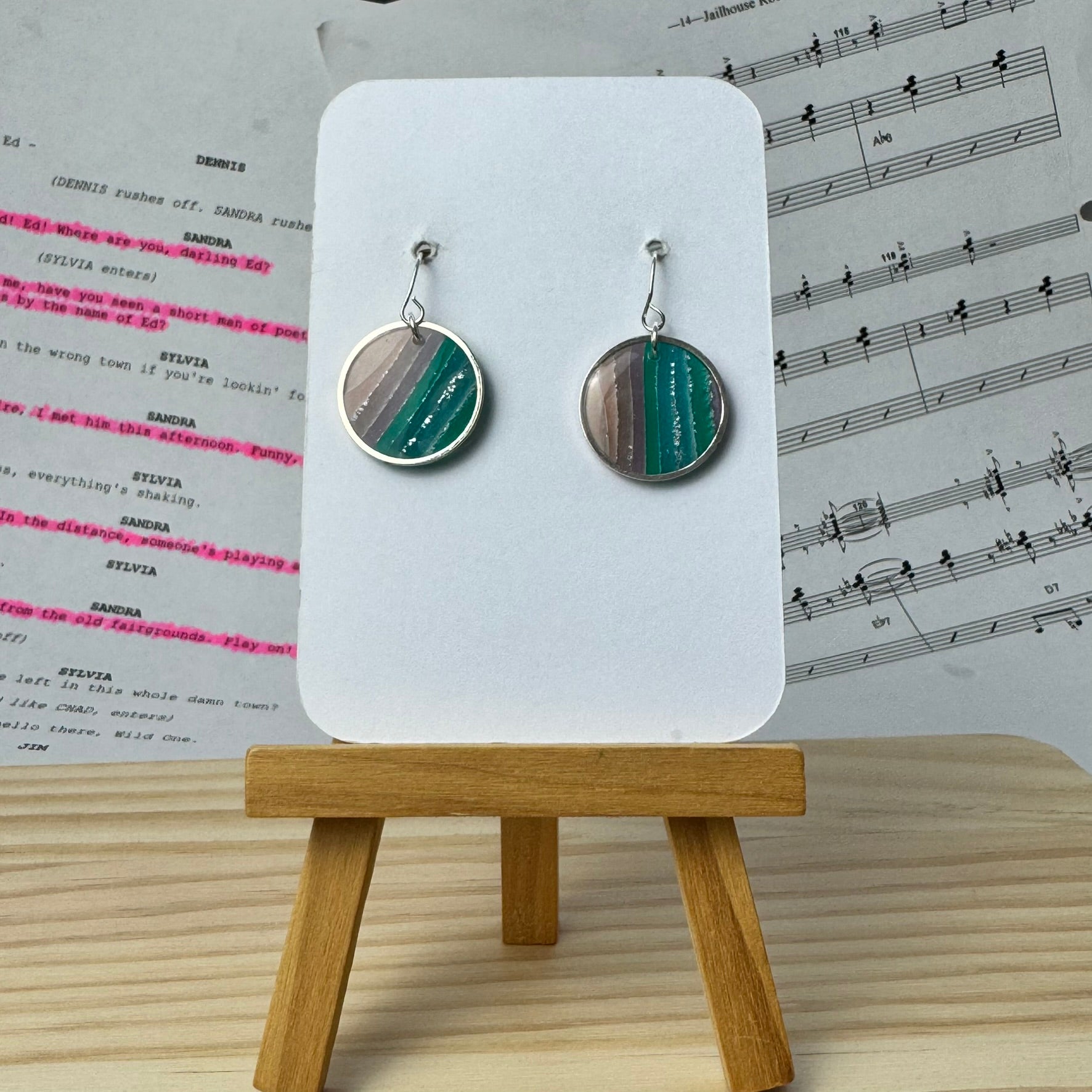 Striped earrings in a silver coloured circle bezel, on a display card with a wooden stand, set against a background of sheet music.