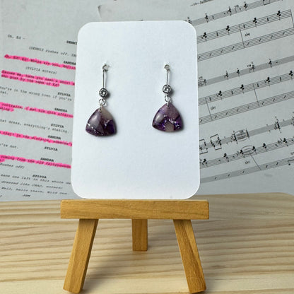 Pair of purple teardrop earrings on a white card with a wooden stand, against a music sheet background.