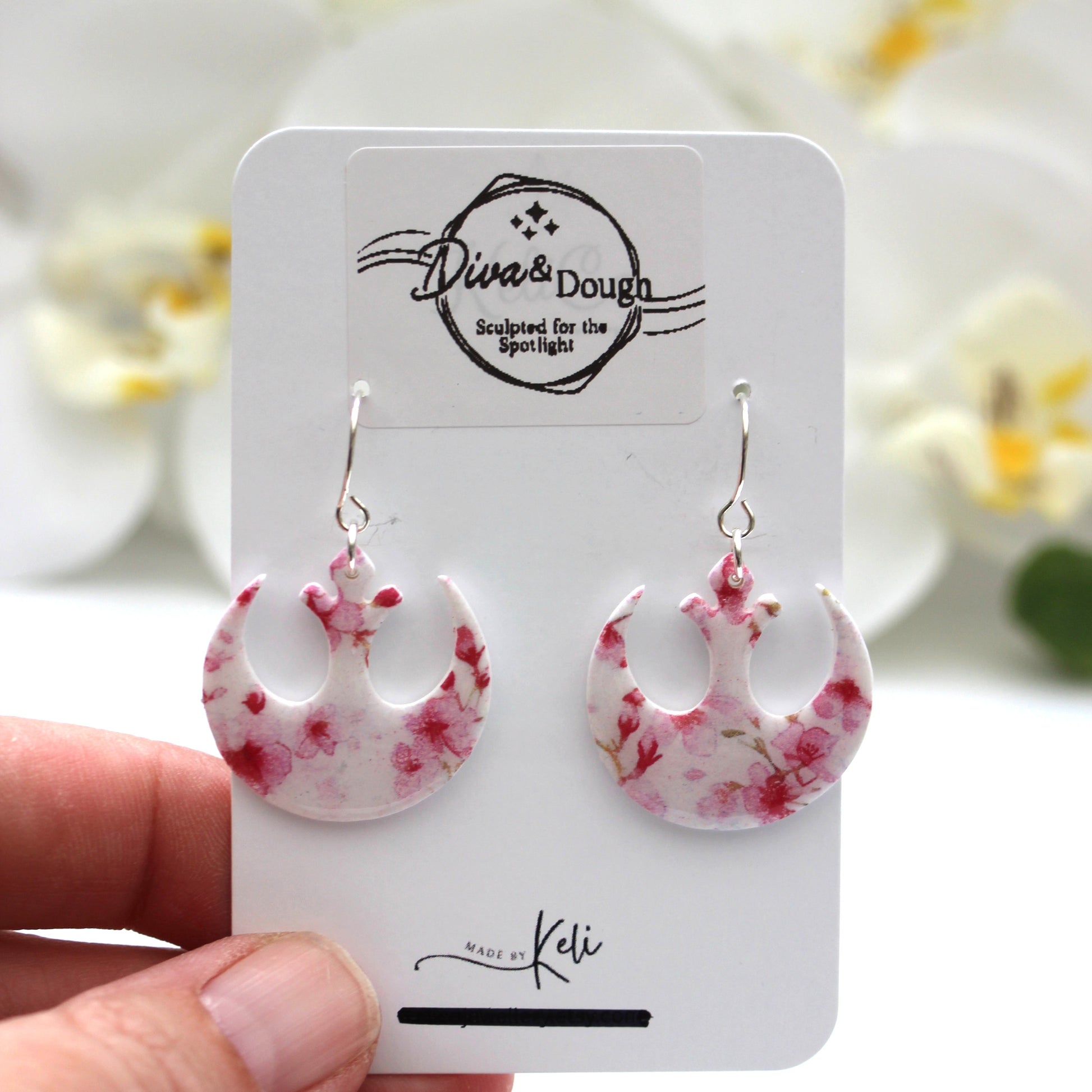 Pink floral earrings on a branded card held by a hand with a blurred floral background