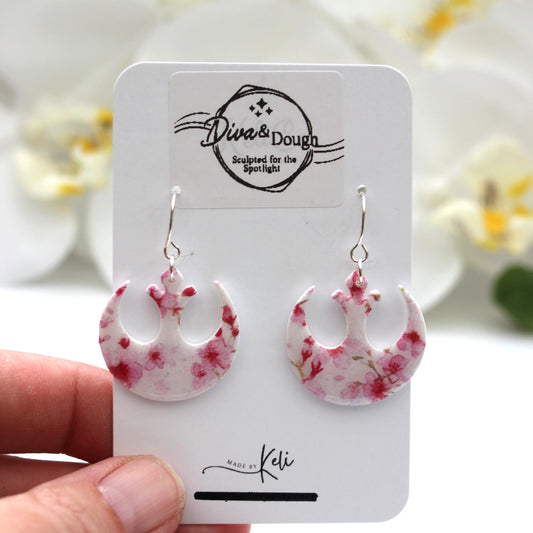 Pink floral earrings on a branded card held by a hand with a blurred floral background