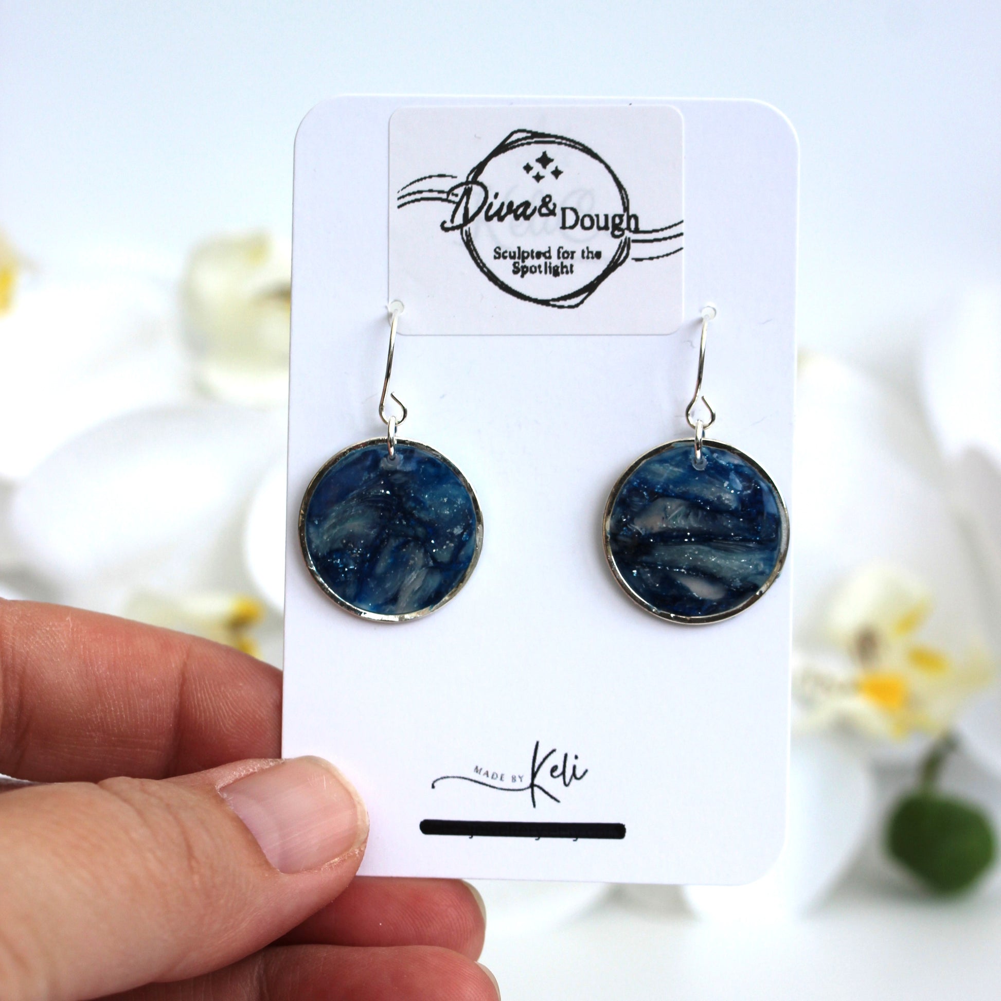 Blue marbled earrings on a branded card held by a hand with a blurred floral background