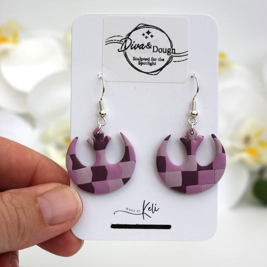 Purple geometric patterned earrings on a branded card held by a hand with a blurred floral background.