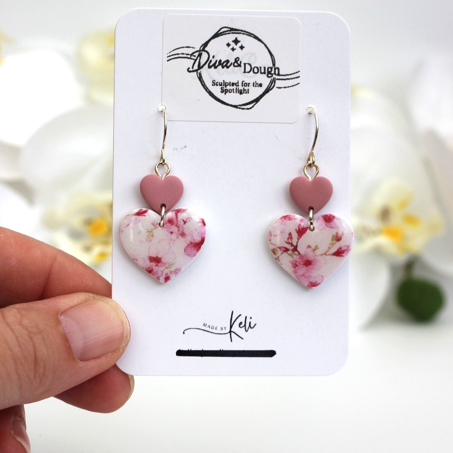 Heart-shaped earrings with a floral print on a branded card held by a hand with a blurred floral background
