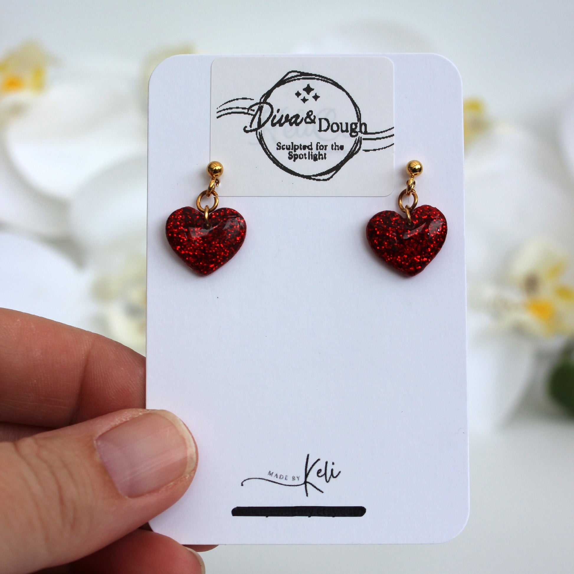 Red heart-shaped earrings on a white card held by a hand with a blurred floral background