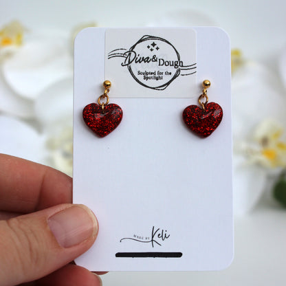 Red heart-shaped earrings on a white card held by a hand with a blurred floral background