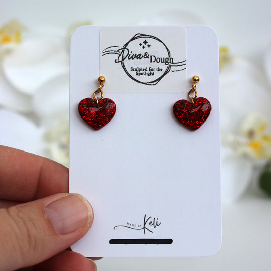 Red heart-shaped earrings on a white card held by a hand with a blurred floral background