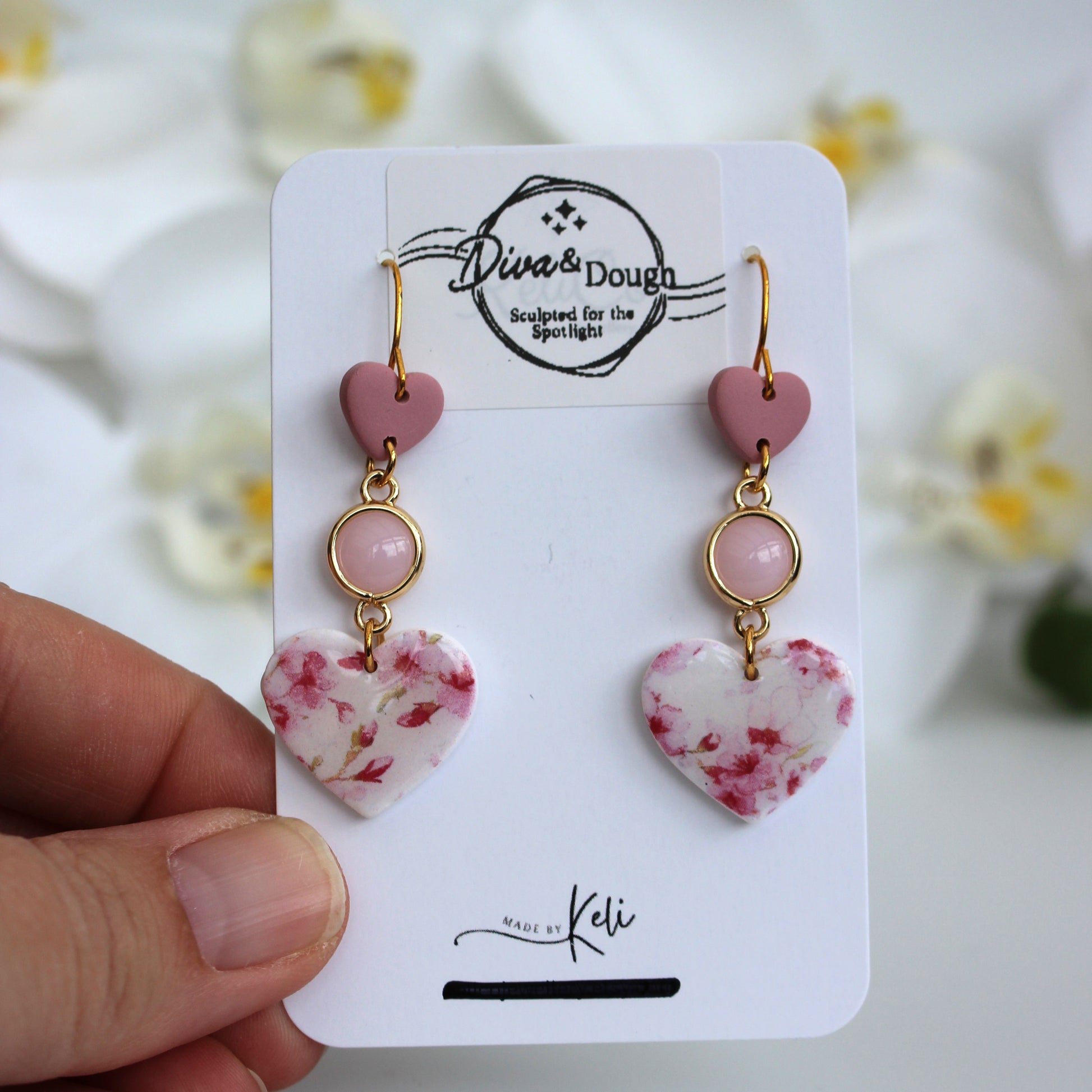 Heart-shaped earrings on a 'Diva & Dough' branded card with a blurred floral background.