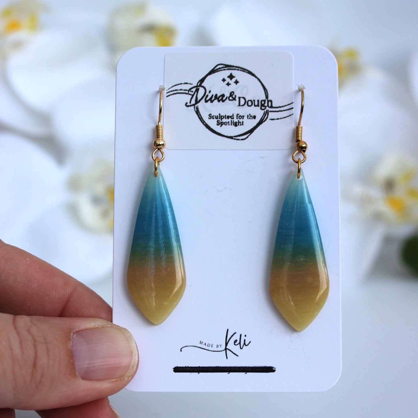 Teal and gold teardrop earrings on a branded card held by a hand with a blurred floral background.