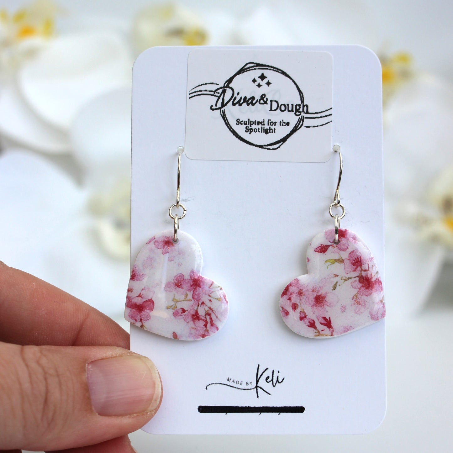 Heart-shaped floral earrings on a branded card held by a hand with a blurred floral background.
