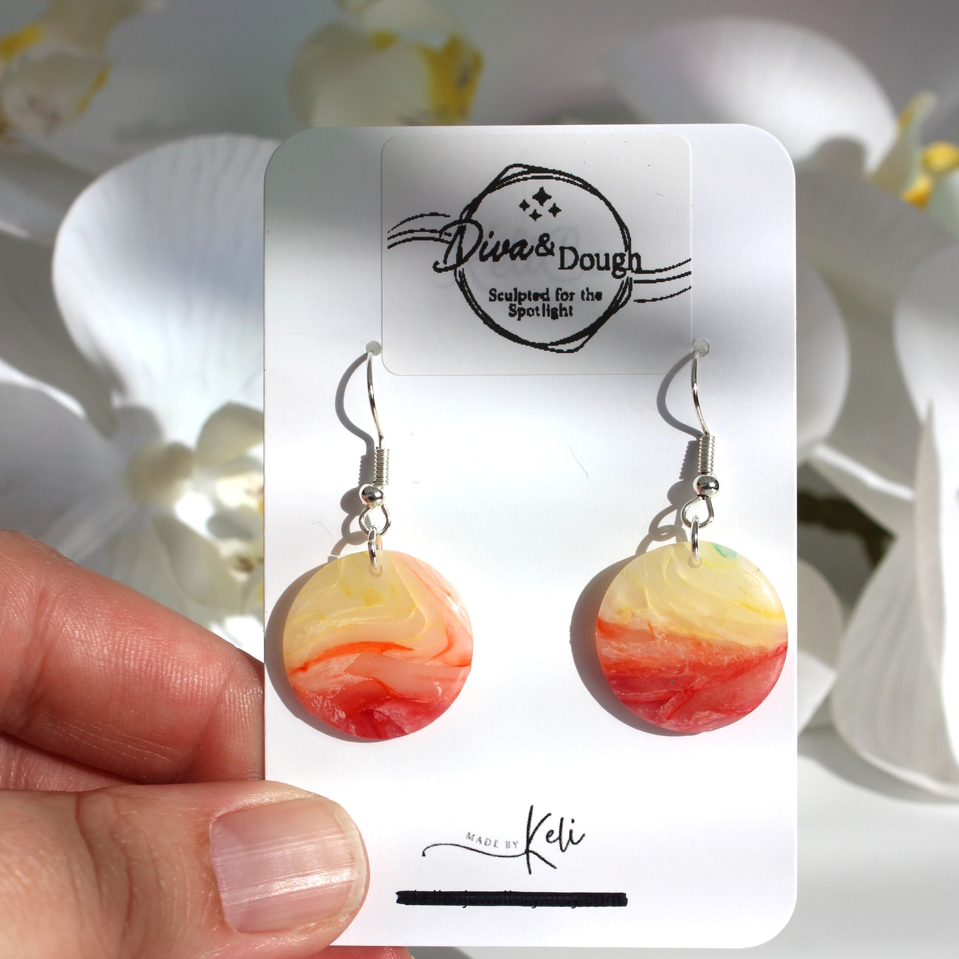 Marbled orange and yellow earrings on a 'Diva & Dough' branded card held by a hand with flowers in the background.