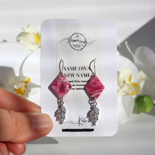 Pink earrings with a crystal accent on a white card held by a hand with flowers in the background
