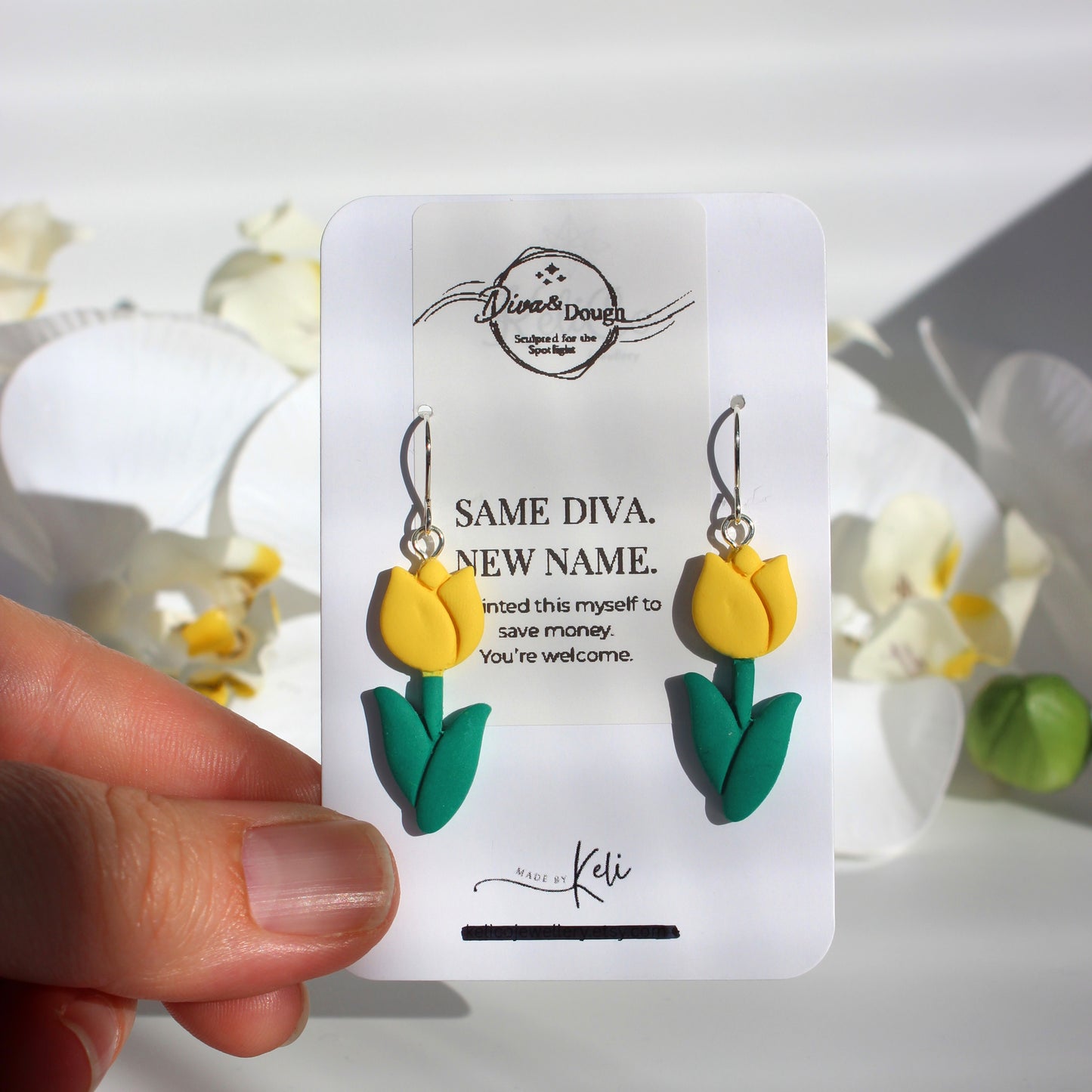 Yellow tulip-shaped earrings on a branded card held by a hand with flowers in the background