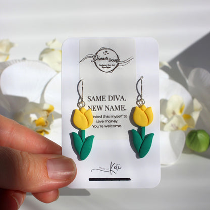 Yellow tulip-shaped earrings on a branded card held by a hand with flowers in the background
