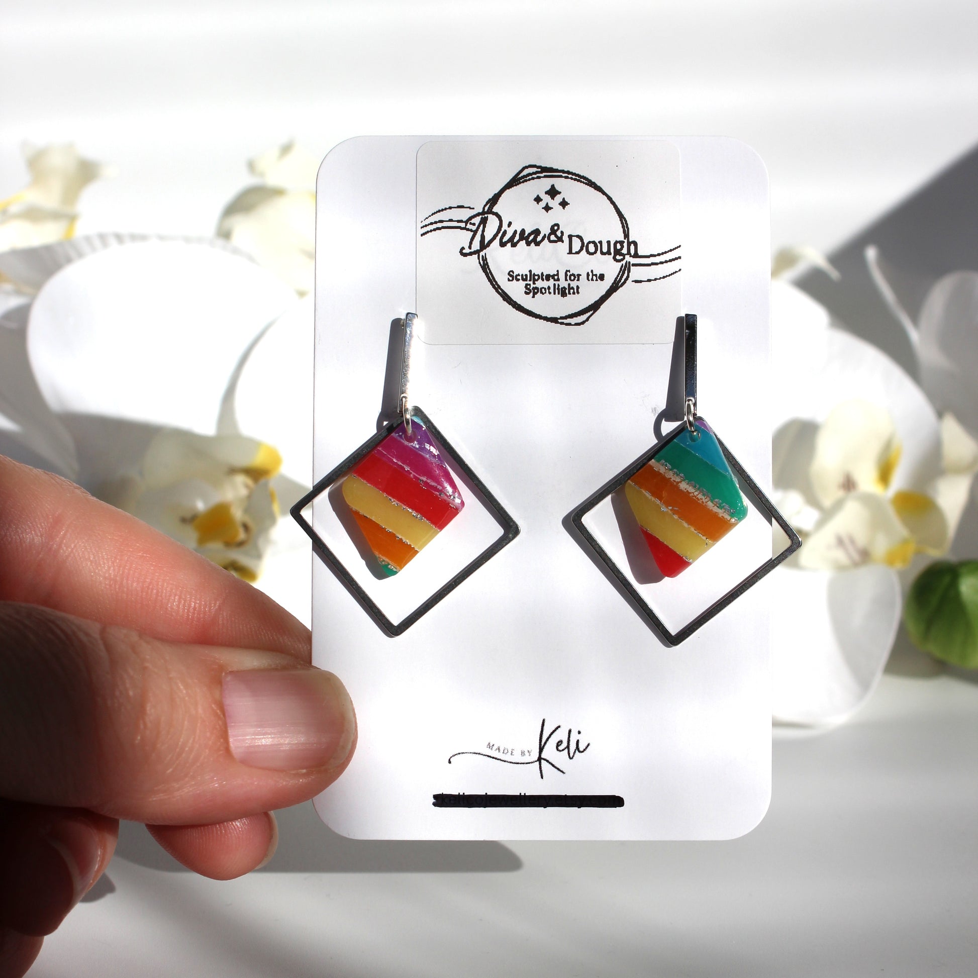 Colorful square earrings on a branded card held by a hand with a blurred floral background