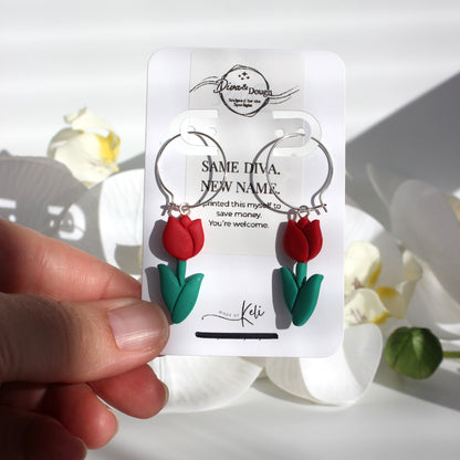 Red tulip earrings on a branded card held by a hand with flowers in the background