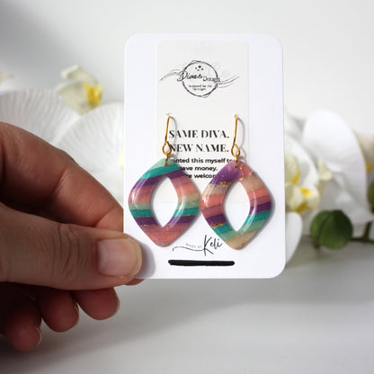 Rainbow hoop earrings on a white card held by a hand with a blurred floral background