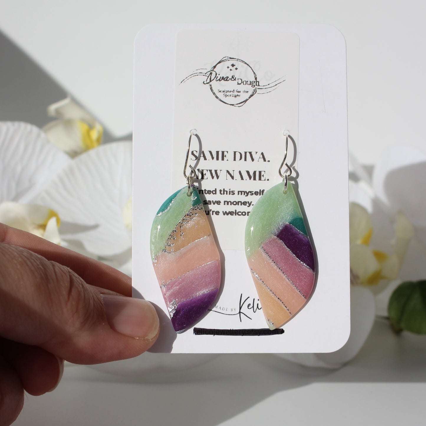 Colorful leaf drop earrings on a branded card held by a hand with a white background.