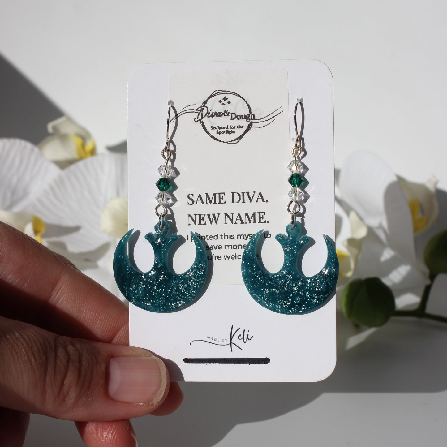 Teal glittery earrings on a white card held by a hand with flowers in the background