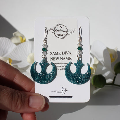 Teal glittery earrings on a white card held by a hand with flowers in the background
