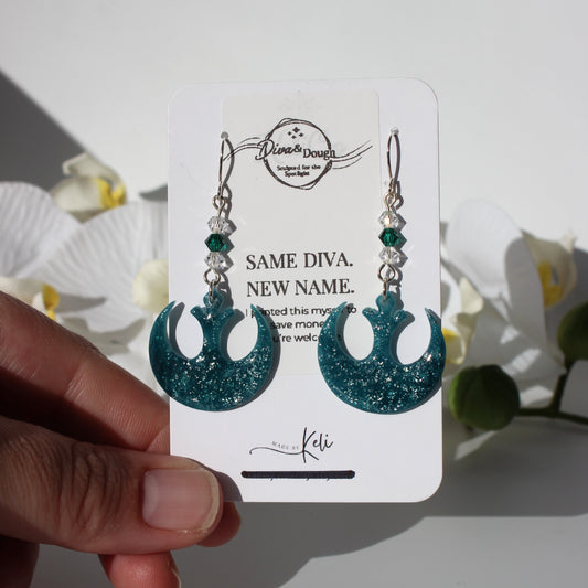 Teal glittery earrings on a white card held by a hand with flowers in the background