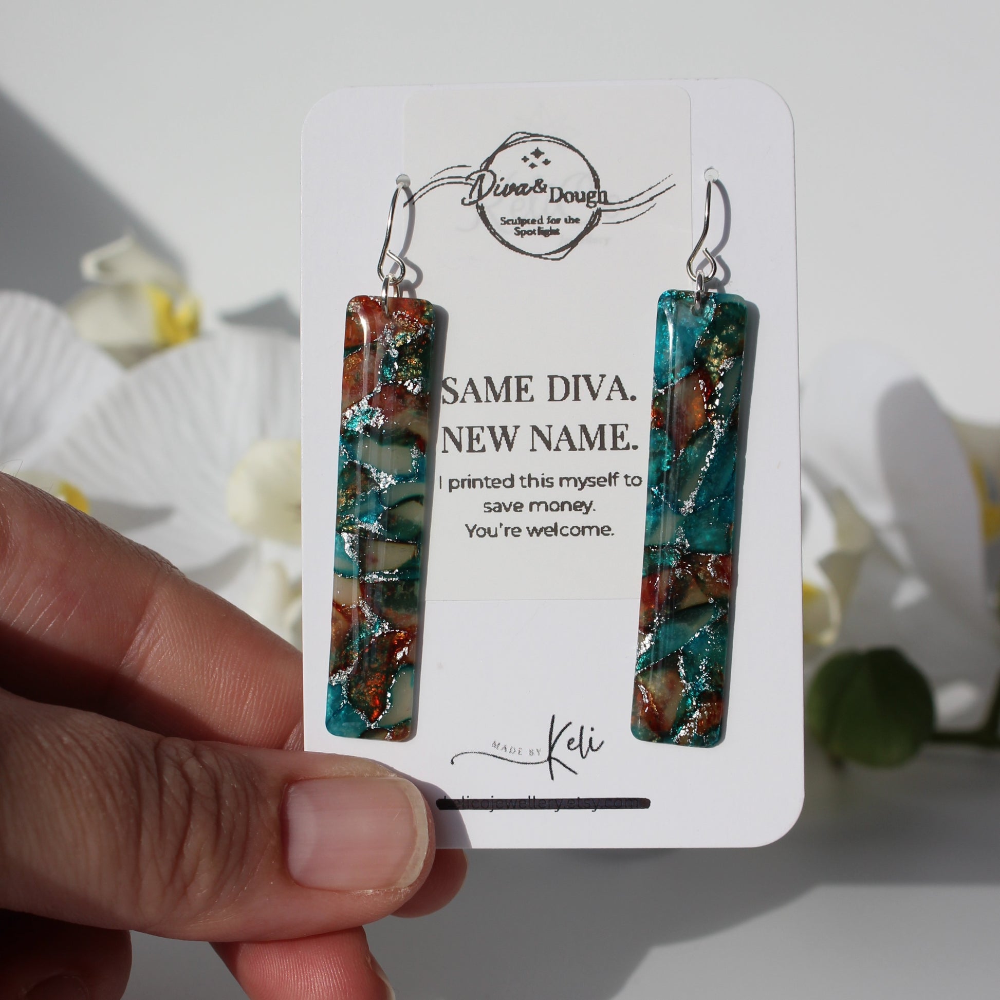 Teal and brown marbled earrings on a branded card held by a hand with a blurred floral background