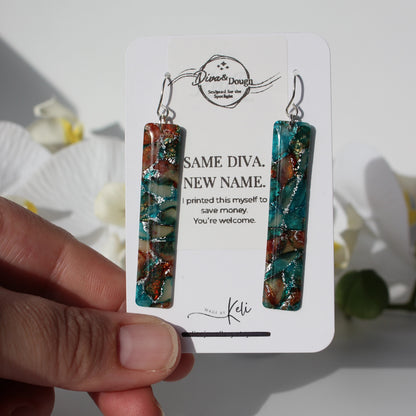 Teal and brown marbled earrings on a branded card held by a hand with a blurred floral background