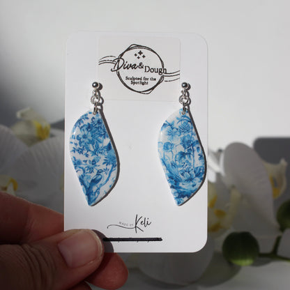 Pair of blue floral earrings on a branded card held by a hand with a blurred floral background.