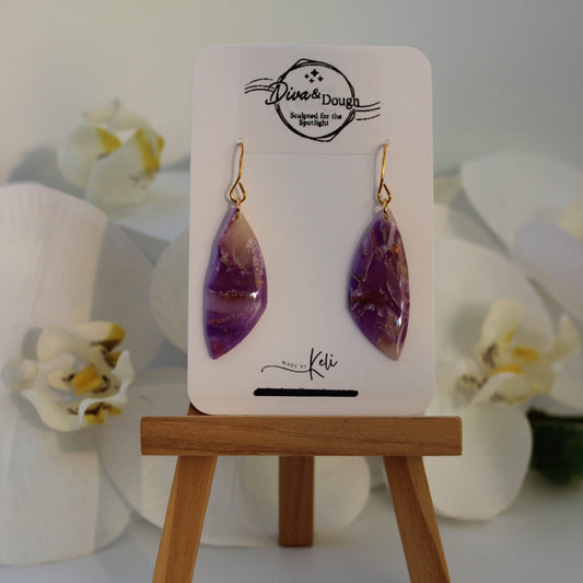 Purple teardrop earrings on a display card with a wooden stand, surrounded by white flowers.