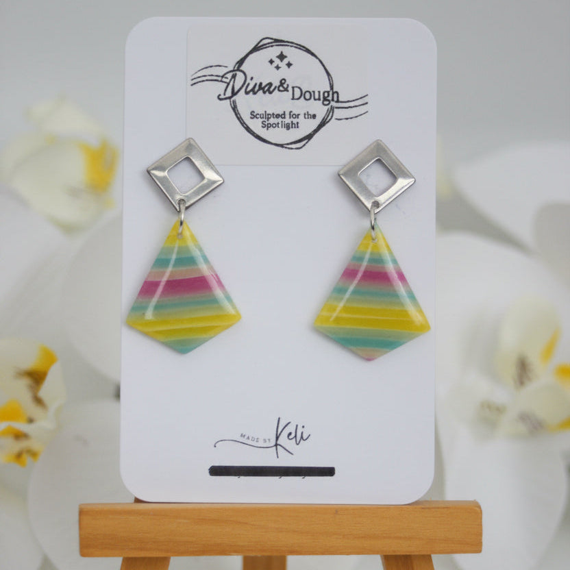 Colorful triangular earrings on a white card with 'Diva & Dough' branding, placed on a wooden stand.
