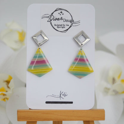 Colorful triangular earrings on a white card with 'Diva & Dough' branding, placed on a wooden stand.