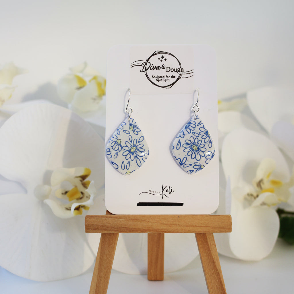 Pair of daisy print earrings on a card with floral design, placed on a white surface with flowers.