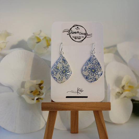 Pair of daisy print earrings on a card with floral design, placed on a white surface with flowers.