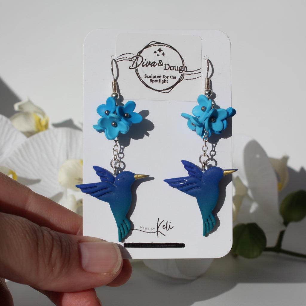 Blue bird-shaped earrings on a branded card held by a hand with a white background.