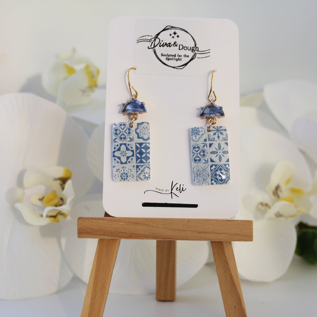 Pair of earrings on a display card with a wooden stand, surrounded by white flowers.
