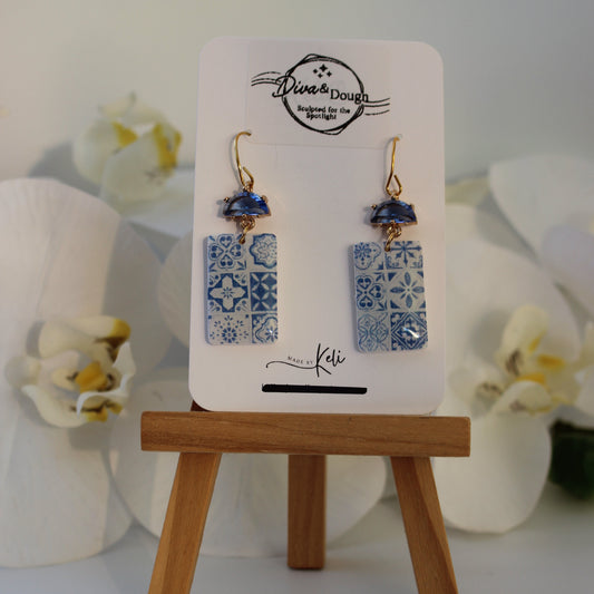 Pair of earrings on a display card with a wooden stand, surrounded by white flowers.