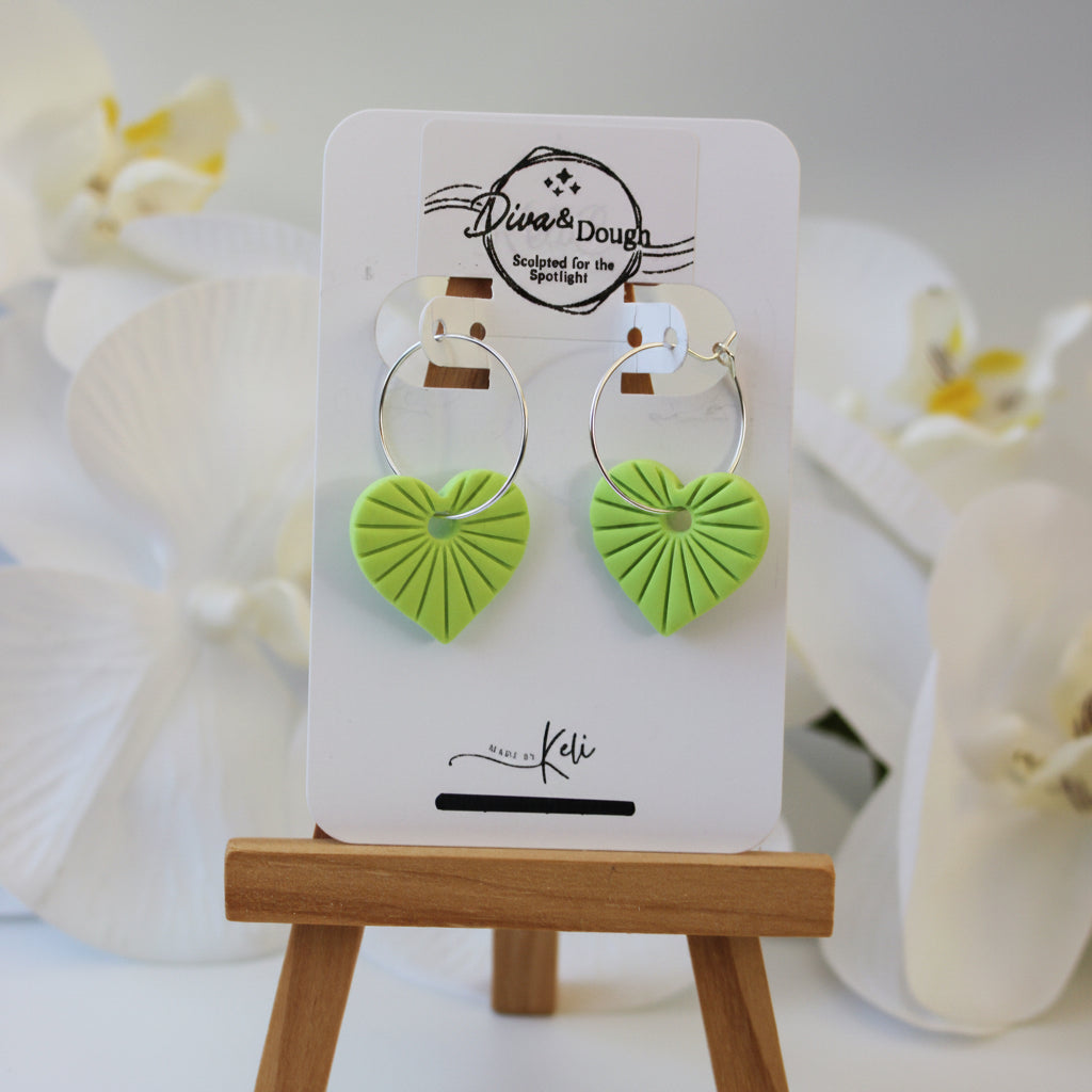 Lime Green heart-shaped earrings on a display card with a wooden stand, white flowers in the background.
