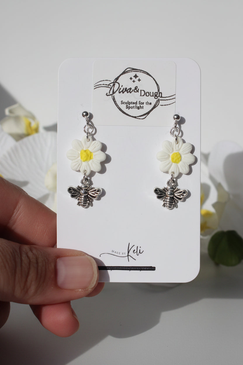Flower-shaped earrings with bee charms on a branded card held by a hand.