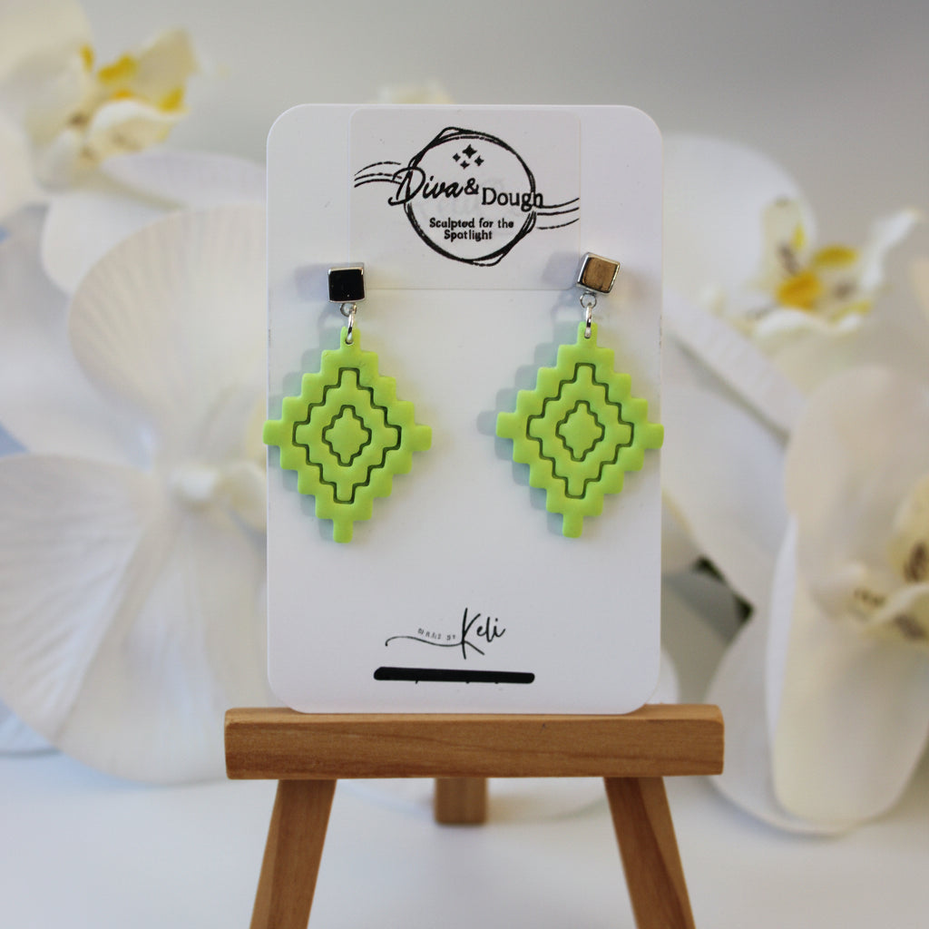 Green geometric earrings on a display card with a wooden stand, white flowers in the background