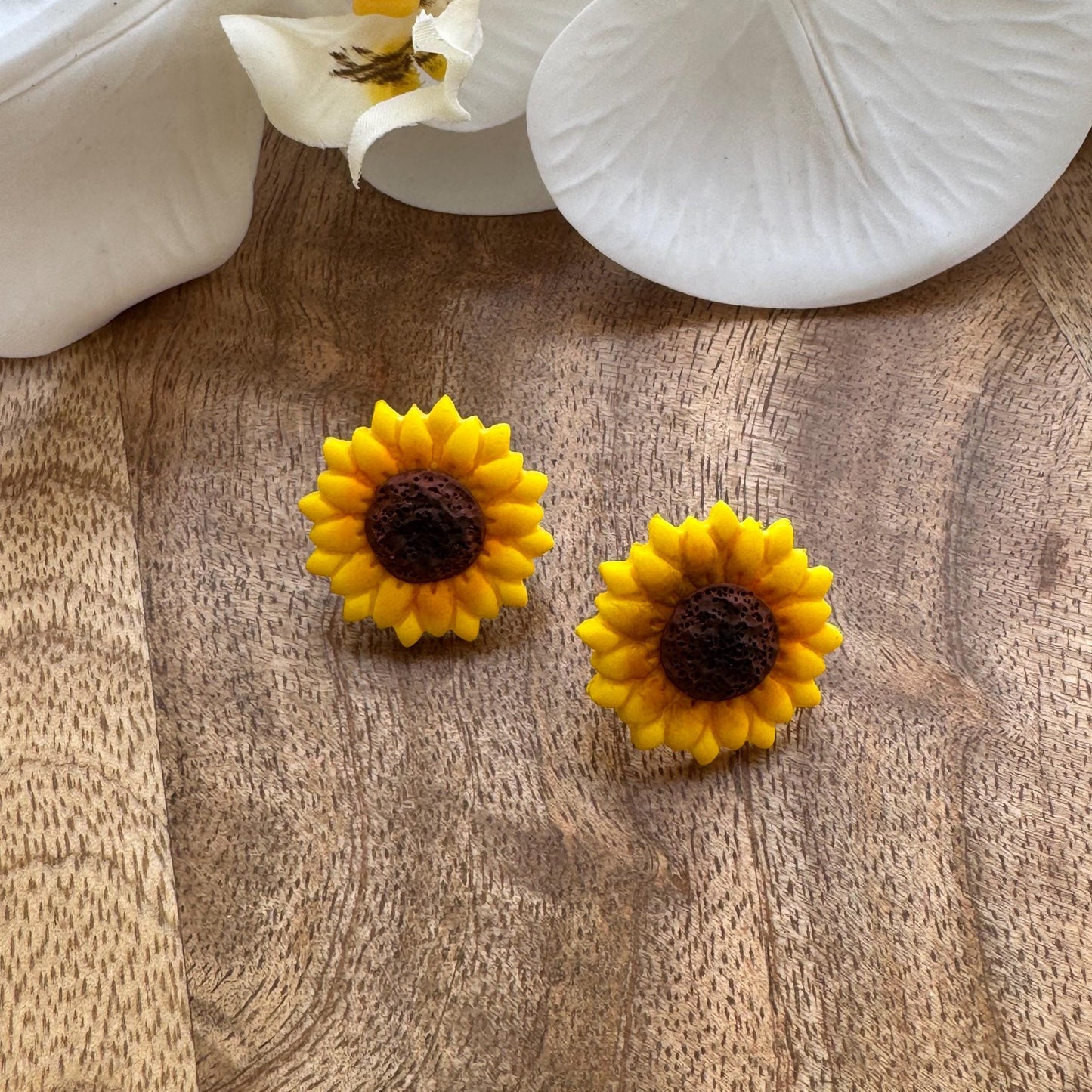 Yellow sunflower-shaped earrings on a wooden surface with white flowers in the background.