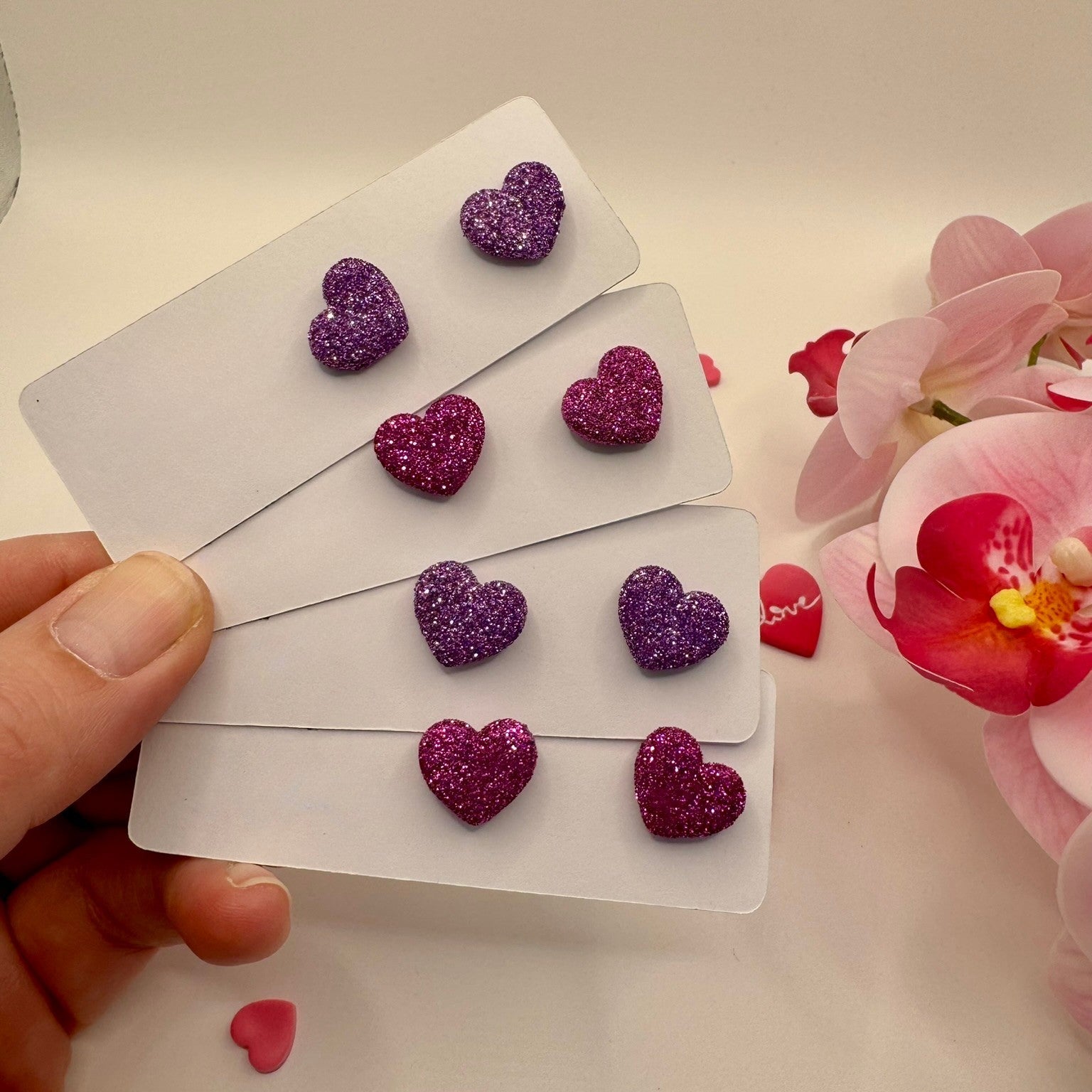 Purple glittery heart-shaped earrings on a card held by a hand with flowers in the background.